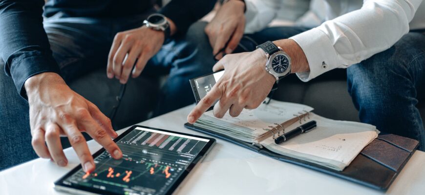 Men in business attire discussing financial data on a tablet indoors.